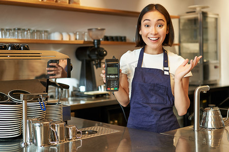 pos机摄影照片_热情的亚洲女咖啡师穿着围裙，展示非接触式信用卡机，看起来对快速非接触式支付感到惊讶，在咖啡店工作