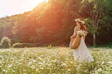 一位身穿白色太阳裙、肩上背着大柳条袋的雏菊花环的年轻女子正穿过一片雏菊田，在森林和夕阳的背景下