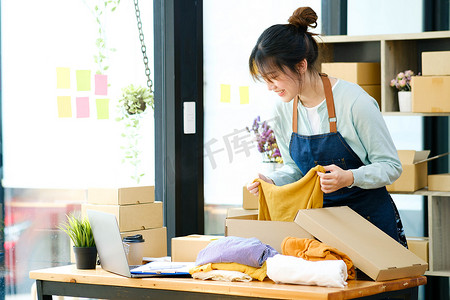 亚洲女服装店老板折叠一件 T 恤，然后用纸板包裹箱包装。