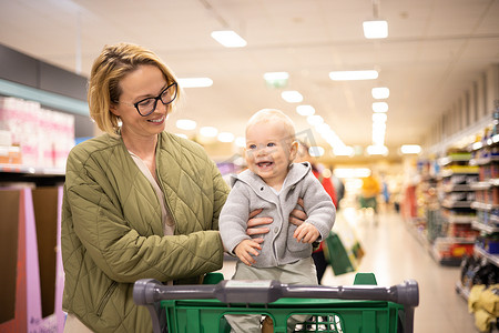 在超市杂货店的部门过道里，妈妈推着购物车带着她的婴儿男婴。