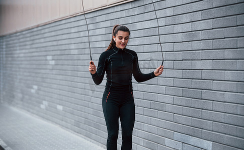 身穿黑色运动服的年轻运动女孩在靠近灰墙的户外用跳绳练习