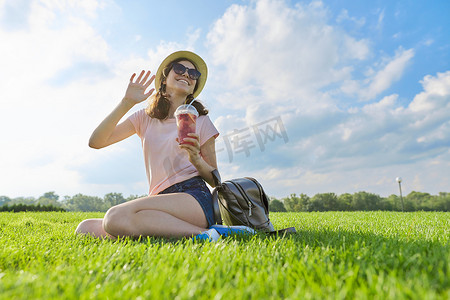 戴帽子、坐在绿草上喝饮料的少女的时尚夏日肖像