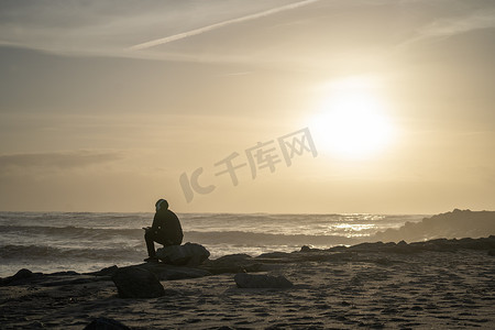 日落时分，男孩戴着耳机坐在海边沙滩上的剪影。 