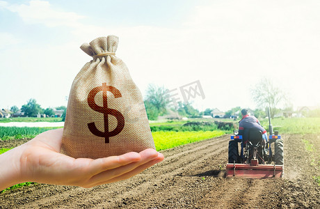 农业补贴摄影照片_一只手在田间耕作拖拉机的背景上伸出一个美元的钱袋。