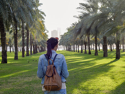 美丽的山水画摄影照片_在阿联酋沙迦的棕榈园里，一位身穿牛仔夹克、背着背包的女性旅行者。