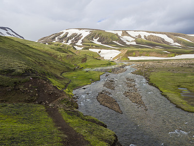 冰岛 f210 路附近地热区的夏季景观，有五颜六色的绿色和橙色苔藓草甸，有温泉和积雪覆盖的流纹岩山脉