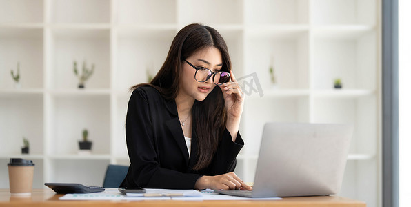 照片中一位美女戴着眼镜坐在办公桌前，周围是办公室的笔记本电脑、文书工作、计算器和咖啡杯。