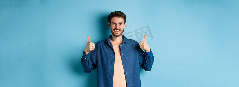 帅气的微笑男人支持，竖起大拇指表示赞同，称赞好工作，在蓝色背景中快乐地站立
