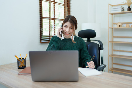 一位亚洲年轻女性的肖像，她在清晨使用办公桌上的电话、电脑和财务文件时面带微笑