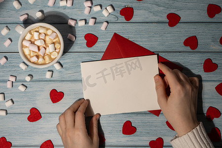 女手拿着空的情人节卡片，在木制的蓝色背景上用白咖啡和棉花糖模拟红包。