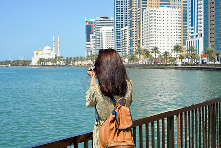 一名身穿长裙、背着背包的女士带着相机沿着沙迦酋长国哈立德湖的 Al Majaz 堤岸行走。