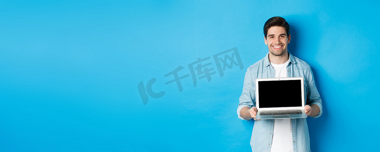 介绍产品摄影照片_帅气的年轻人在笔记本电脑屏幕上介绍产品，展示电脑，微笑，站在蓝色背景上