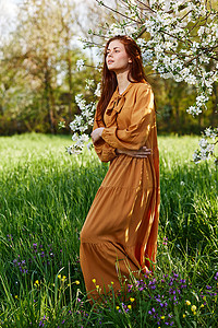 一个苗条、甜美的女人穿着橙色长裙站在一棵开花的树旁高高的草丛中，双臂交叉放在胸前，望向远方