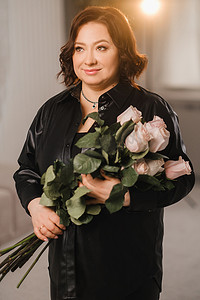 皮衣摄影照片_一位穿着黑色皮衣的时尚成年女性站着，室内有一束粉红玫瑰