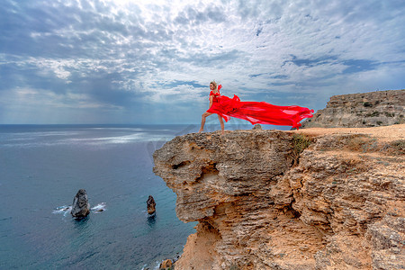 飘动摄影照片_一位身着红色丝绸连衣裙的女子站在海边，背靠群山，她的裙子在微风中摇曳。