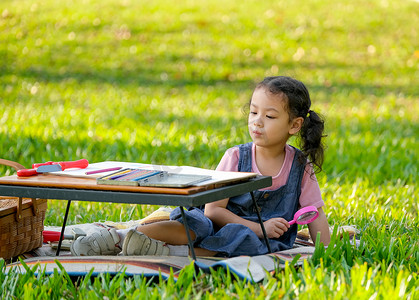 可爱的亚洲小女孩拿着放大镜，在花园里拿着彩色铅笔和玩具坐在桌子旁，晨光.