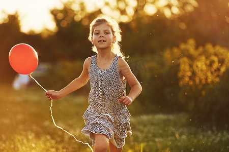 草地玩的孩子摄影照片_夏天，手里拿着红气球的快乐小女孩在草地上玩得开心