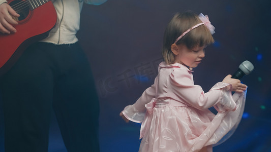 唱歌摄影照片_穿着复古裙子的小女孩在舞台上唱歌，她的父亲弹着原声吉他