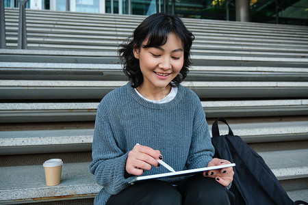 微笑的女孩，平面设计师，使用数字平板电脑和钢笔工具绘画，为大学做家庭作业，坐在楼梯上