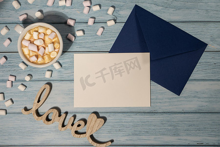 空的问候语或邀请卡模拟了蓝色信封，白色的咖啡和棉花糖在木制的蓝色背景上。