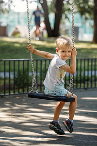 淘气男孩摄影照片_一个淘气的小男孩在公园里荡秋千，孩子转向镜头，开心地笑着
