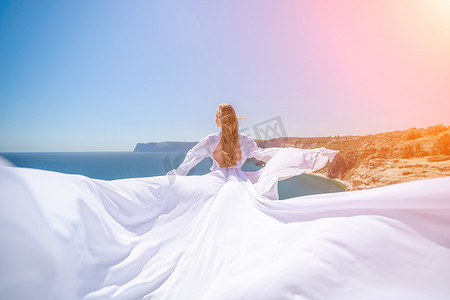 一位长发的金发女子身着飘逸的白色长裙站在阳光明媚的海边，丝绸面料随风飘扬，背景是蓝天和群山。