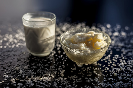 纯牛奶摄影照片_关闭玻璃碗纯牛奶与热牛奶在黑色木质光滑表面上充分混合，以及生酥油澄清黄油和一些糖晶体散布在表面上。