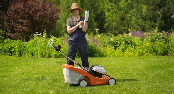 一个戴着草帽的年轻人正在戴上手套，在他美丽的绿色花卉夏季花园里用割草机修剪草坪。