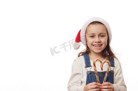 戴着圣诞帽的漂亮小女孩，拿着心形的圣诞甜棒棒糖，微笑着看着镜头