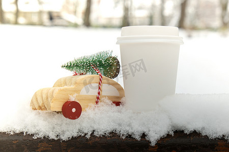 木制汽车载着圣诞树，纸杯在雪地上模拟咖啡或热巧克力。