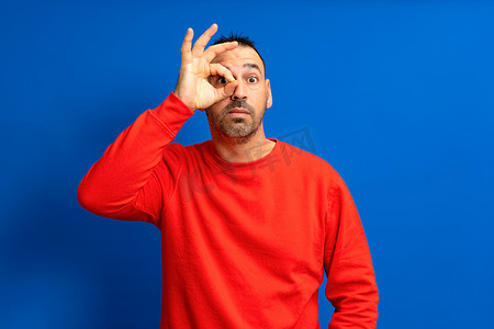 40 多岁的留着胡子的西班牙裔男子身穿红色套头衫，在蓝色工作室背景下，一只手用望远镜观察。