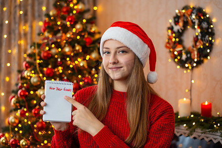 新年愿望清单摄影照片_戴着红色圣诞帽的年轻美女，用手指指着题词的新年目标