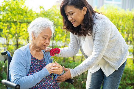 Caregver 帮助亚洲老年妇女拿着红玫瑰花，在阳光明媚的花园里微笑和快乐。