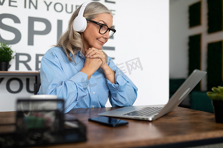 50 岁的女经理在在线会议期间戴着耳机坐在桌子旁看着笔记本电脑。 