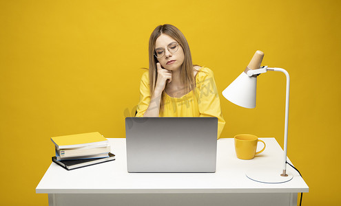 年轻成功的秘书雇员女商人的工作室肖像穿着黄色 T 恤坐在白色办公桌前工作，笔记本电脑在线浏览互联网隔离在黄色背景。