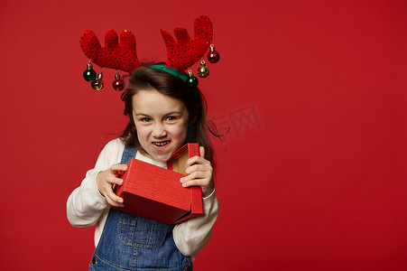 穿着鹿角圈的调皮风趣的小女孩，打开圣诞惊喜礼盒，红色背景