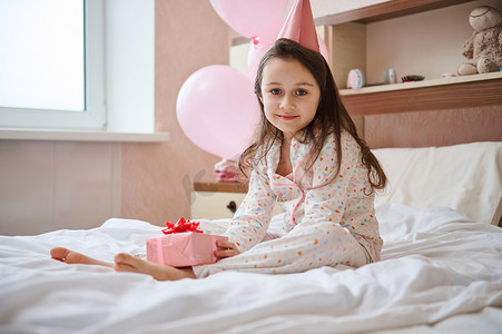 拿着礼盒摄影照片_戴着粉色节日帽、拿着礼盒、坐在卧室床上、拿着氦气球的可爱生日女孩