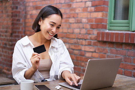 微笑的亚洲女性坐在咖啡馆里，拿着笔记本电脑，用信用卡支付网上购物费用，发送她的银行账户详细信息