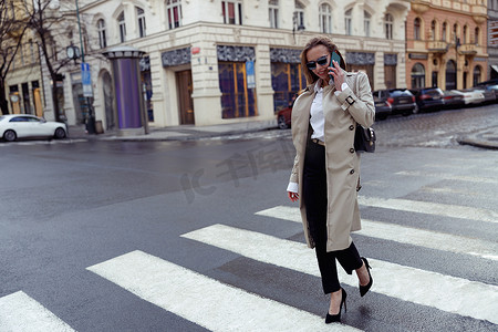 穿着时髦服装的女性在城市建筑的背景下过马路并打电话
