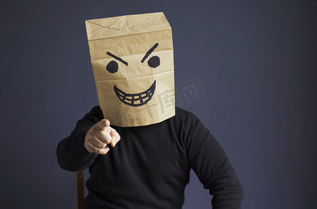 一个身穿黑色高领毛衣的男人头上拿着一个纸袋，上面有愤怒的表情符号，他的手指向前方。