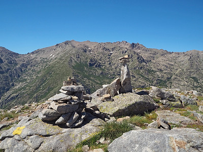 石凯摄影照片_科西嘉阿尔卑斯山的高山风景，有绿色灌木和岩石桩石标，在 gr20 跋涉和白云蓝天上标有路径