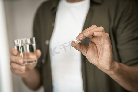 男人一只手拿着药丸，另一只手拿着一杯水的特写镜头，在他家的舒适环境中。