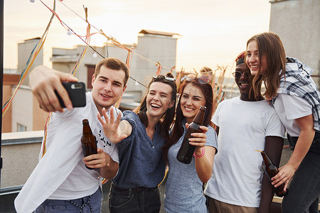 年轻人年轻人聚会摄影照片_一群穿着休闲服的年轻人白天在屋顶一起聚会，并通过电话自拍