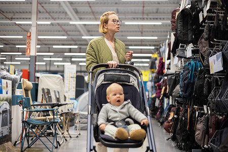 穿着随意的母亲在超市商店的运动部门选择运动鞋和服装产品，她的婴儿男婴坐在婴儿车里。