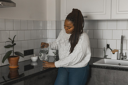 口渴的非洲裔美国妇女在厨房里喝矿泉水，从水壶里倒健康的液体到玻璃杯里，带辫子的快乐女士享受清爽的饮料