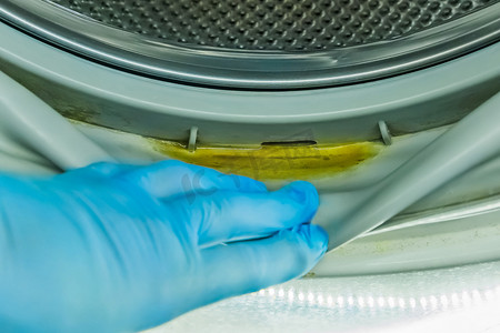 洗衣机洗衣机摄影照片_一个戴着橡胶家用手套的男人的手指向洗衣机旧滚筒压实机中的脏水残留