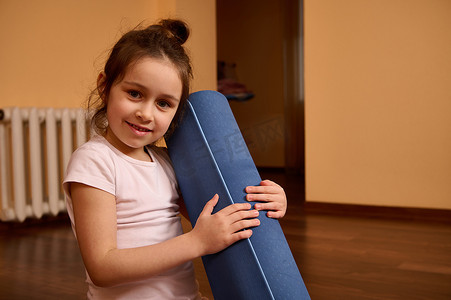 特写：一个微笑的小女孩拿着健身垫，看着相机，在室内享受锻炼和健身