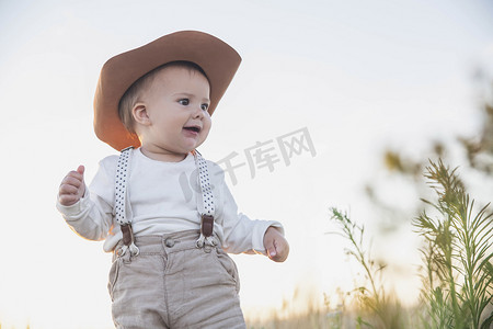 戴着帽子和吊带的婴儿在日落时看着田野