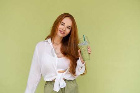 身着绿色背景休闲衬衫的漂亮红发女性在炎热的夏日享受新鲜的冰绿抹茶和牛奶