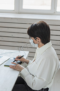 医用面罩平面设计师或摄影师在现代办公室工作时使用数字图形平板电脑的中年妇女，坐在现代工作区的专业女性修饰师，通用设计平板电脑和附近的相机。 